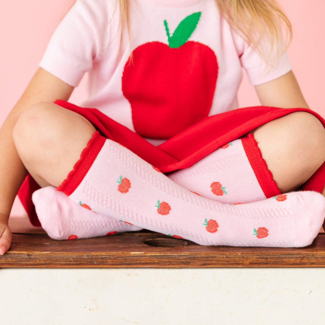 girls sitting criss cross on desk in pink knee high socks with red apple design and red scalloped trim and matching red skirt and apple sweater