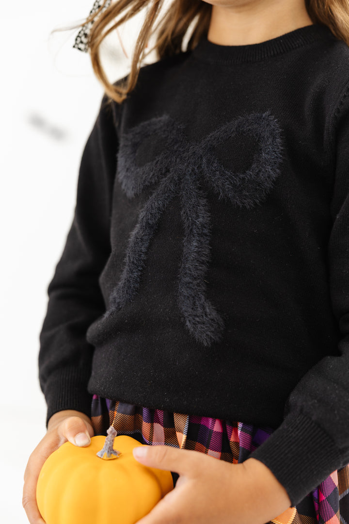 closeup of fluffy black bow detail for black knit Halloween sweater on little girl holding mini pumpkin