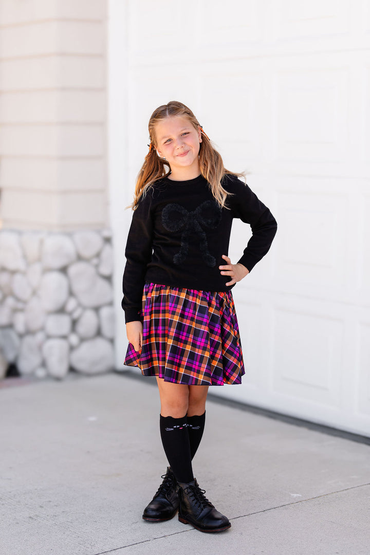 little girl in black pullover sweater with fluffy bow design paired with plaid skort in Halloween colors and black cat knee high socks