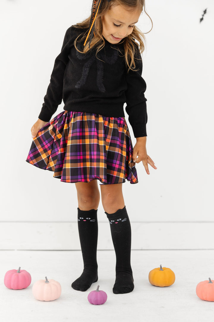 young girl with colorful pumpkins in black bow sweater, Halloween plaid skort and matching black cart knee highs