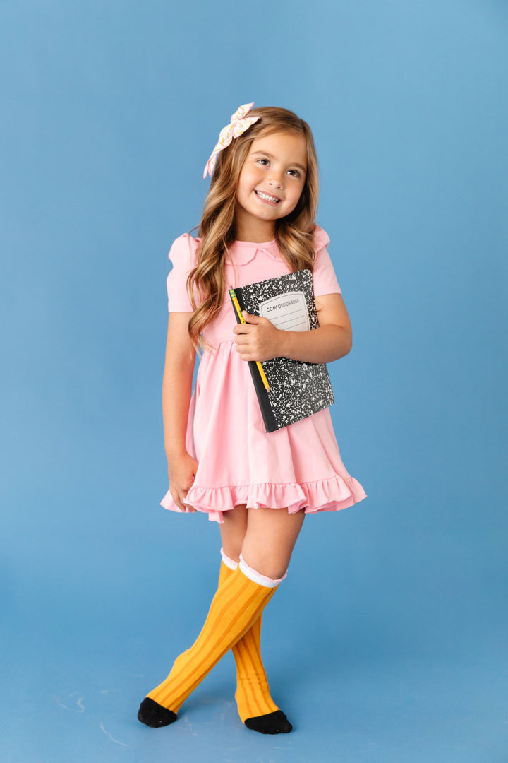 little girl holding composition book in pink collared twirl dress, pencil print hair bow and matching pencil inspired knee high socks