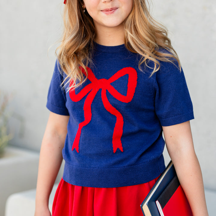 little girl holding schoolbooks in navy blue short sleeve cotton knit sweater with red bow design paired with matching red skort
