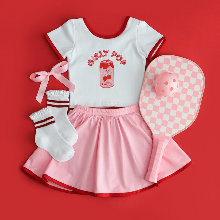 sporty summer flatlay of girls fitted white t-shirt with red binding and "Girly Pop" soda print paired with matching pink skort and summer accessories