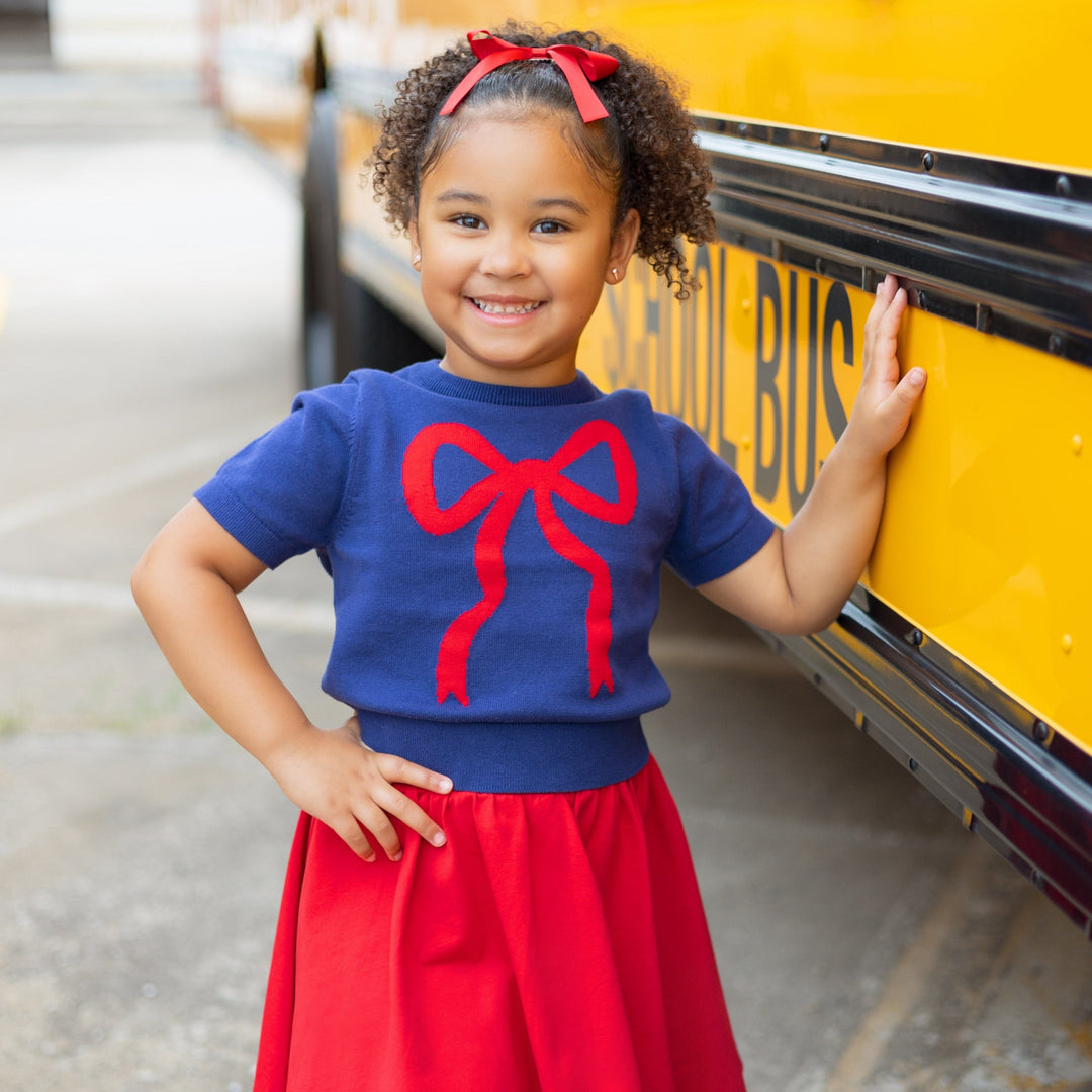 girl standing by school bus in navy short sleeve cotton sweater with red bow on it and matching red skort and satin bow