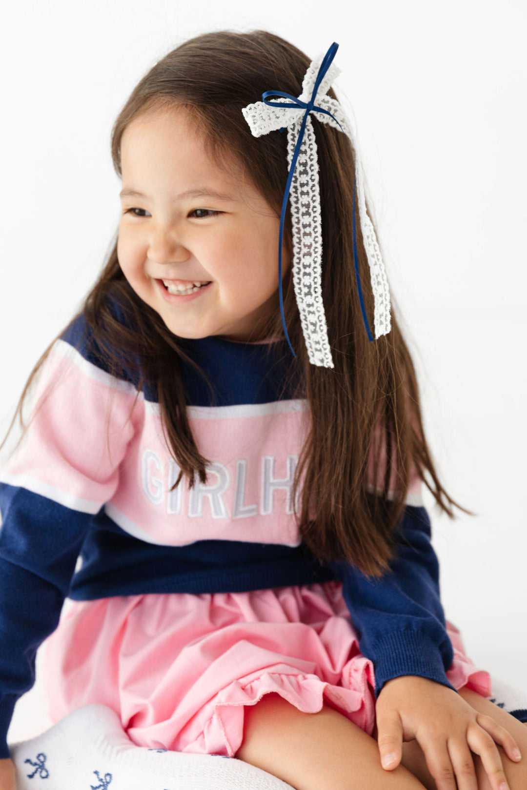 little girl sitting in navy and pink cotton knit pullover with embroidered patches spelling girlhood paired with pink dress, lace ribbon bow and matching navy bow knee highs