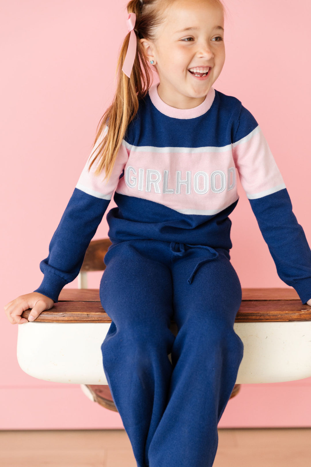 little girl sitting on school desk in navy and pink "girlhood" pullover sweater and matching navy blue sweater pants