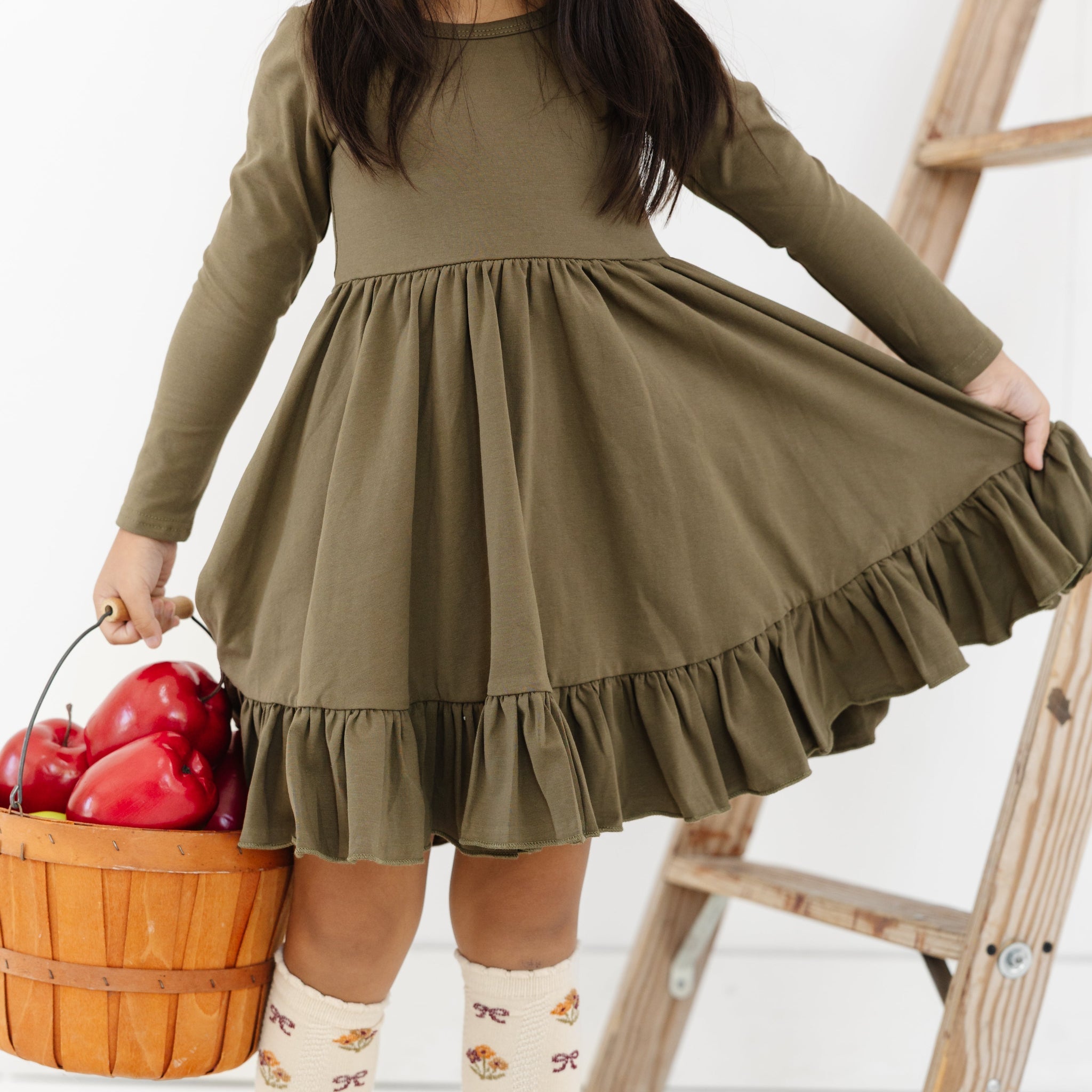 little girl holding basket of apples in olive green cotton blend twirl dress with ruffle hem and coordinating cream scallop trimmed knee high socks with flower and bow print