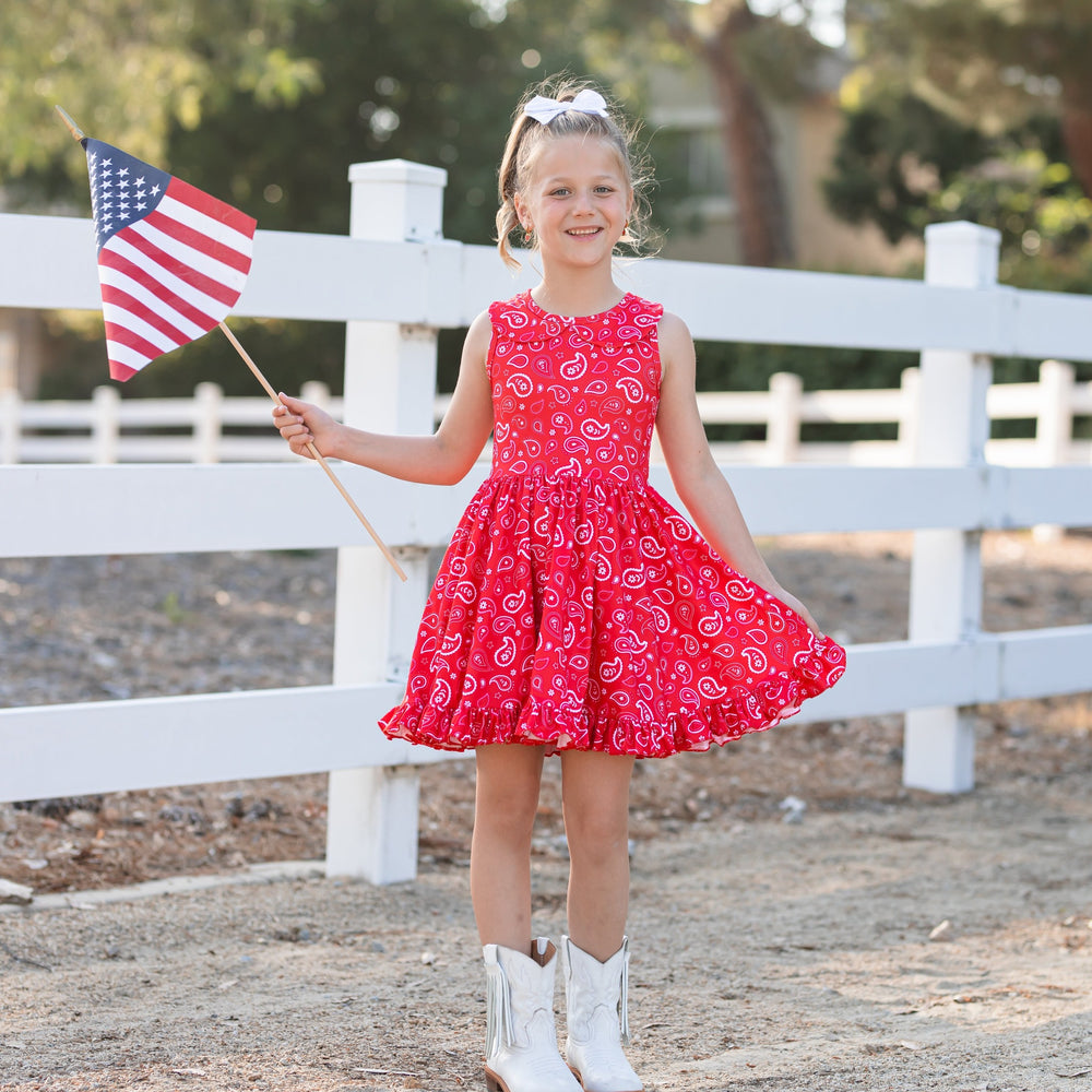 little girl holding american flag wearing red and white paisley print twirl dress and cowgirl boots