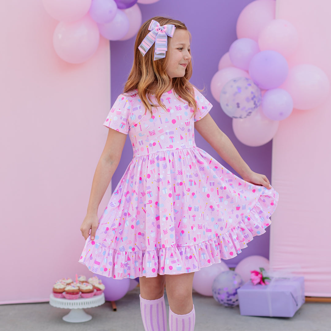 girl in birthday outfit of pink twirl dress with presents and balloons on it and matching purple striped knee highs and pink and purple striped double bow