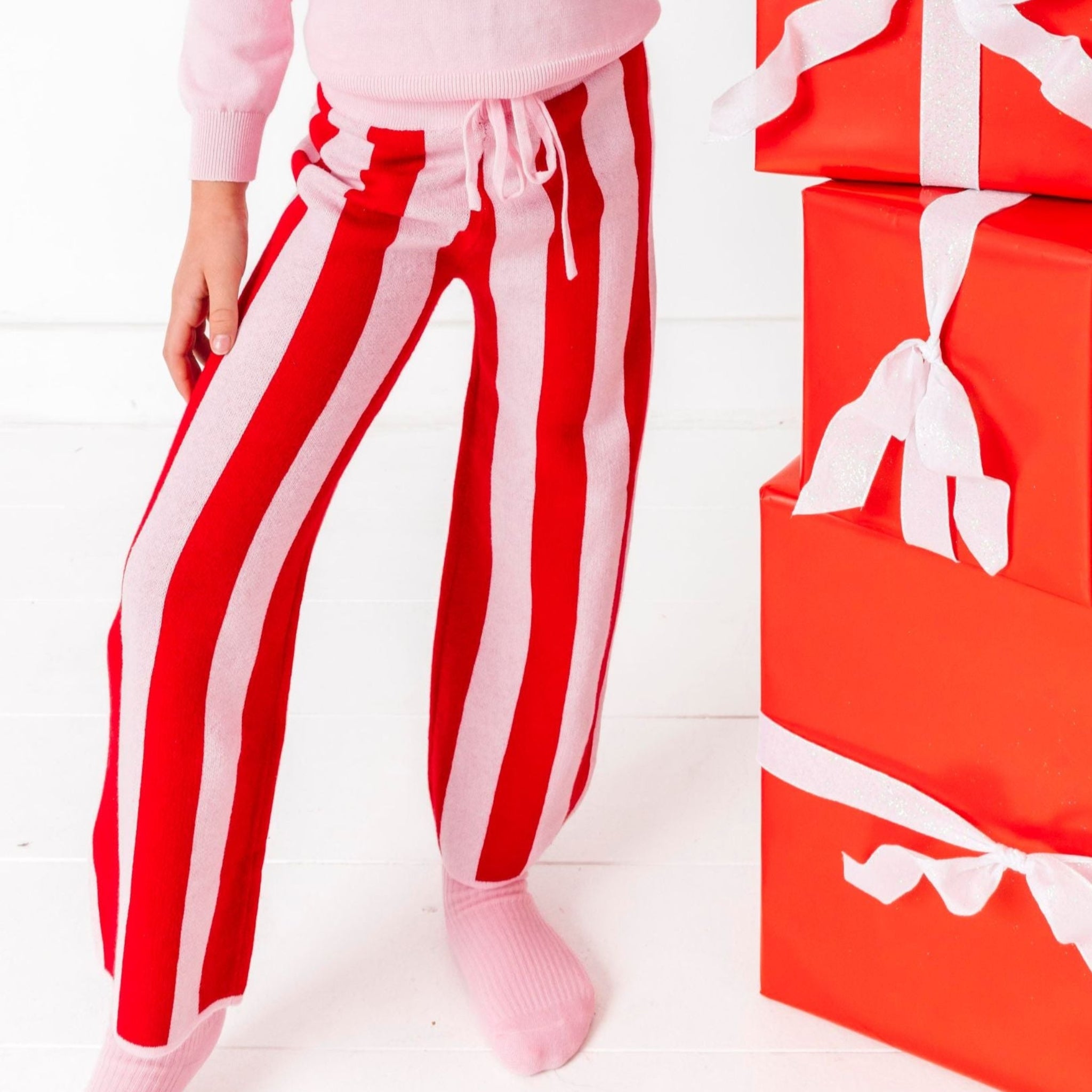 girl by stack of Christmas gifts wearinfgpink pullover sweater with candy cane design and matching red and pink striped sweater pants