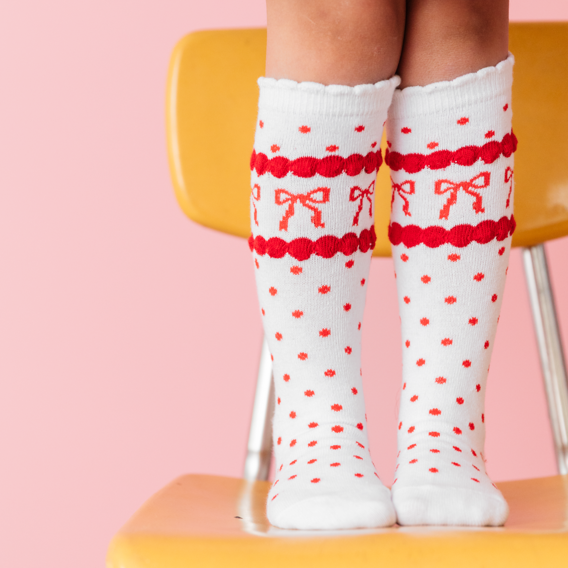 girl wearing scallop trimmed knee highs in white with red dots and red bow print standing on yellow school chair