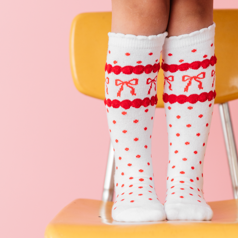 girl wearing scallop trimmed knee highs in white with red dots and red bow print standing on yellow school chair
