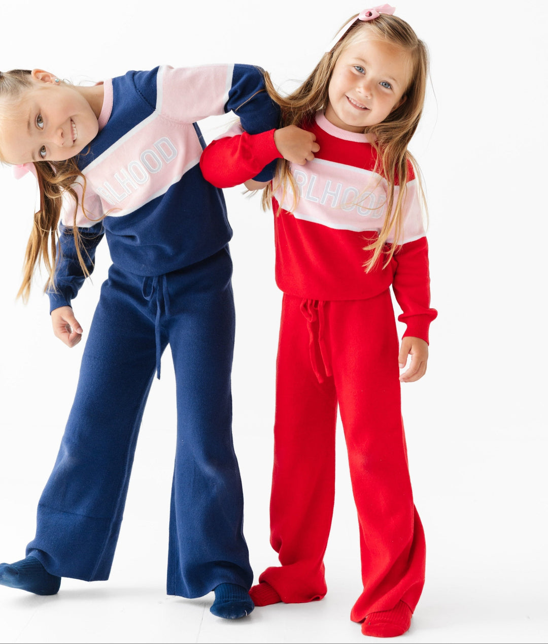 sisters wearing matching cotton knit outfits, one navy and pink and one red and pink with girlhood varsity sweater and solid pants