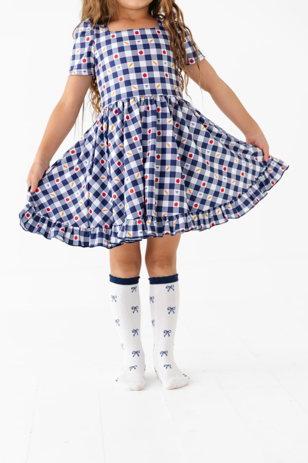 girl holding out skirt of short sleeve, square neck back to school dress in navy and white gingham with apple, pencil and bow print details