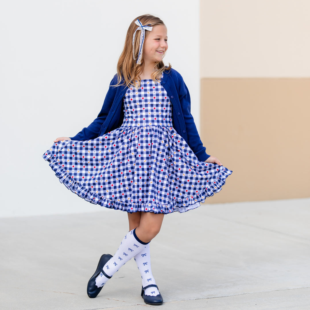 girl in short sleeve, square neck back to school dress in navy and white gingham with apple, pencil and bow print details with navy cardigan sweater and matching bow knee high socks