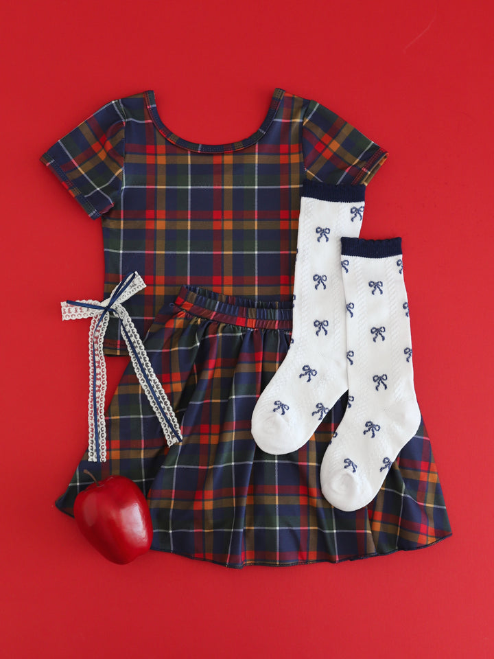 styled flat lay of little girl's back to school outfit featuring matching navy plaid t-shirt and skort paired with white with navy blue bow pattern socks and matching lace ribbon bow