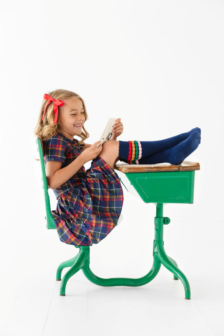 little girl sitting at school desk in matching navy, red and mustard plaid tee-shirt and skort with red satin bow and navy bubble knee highs