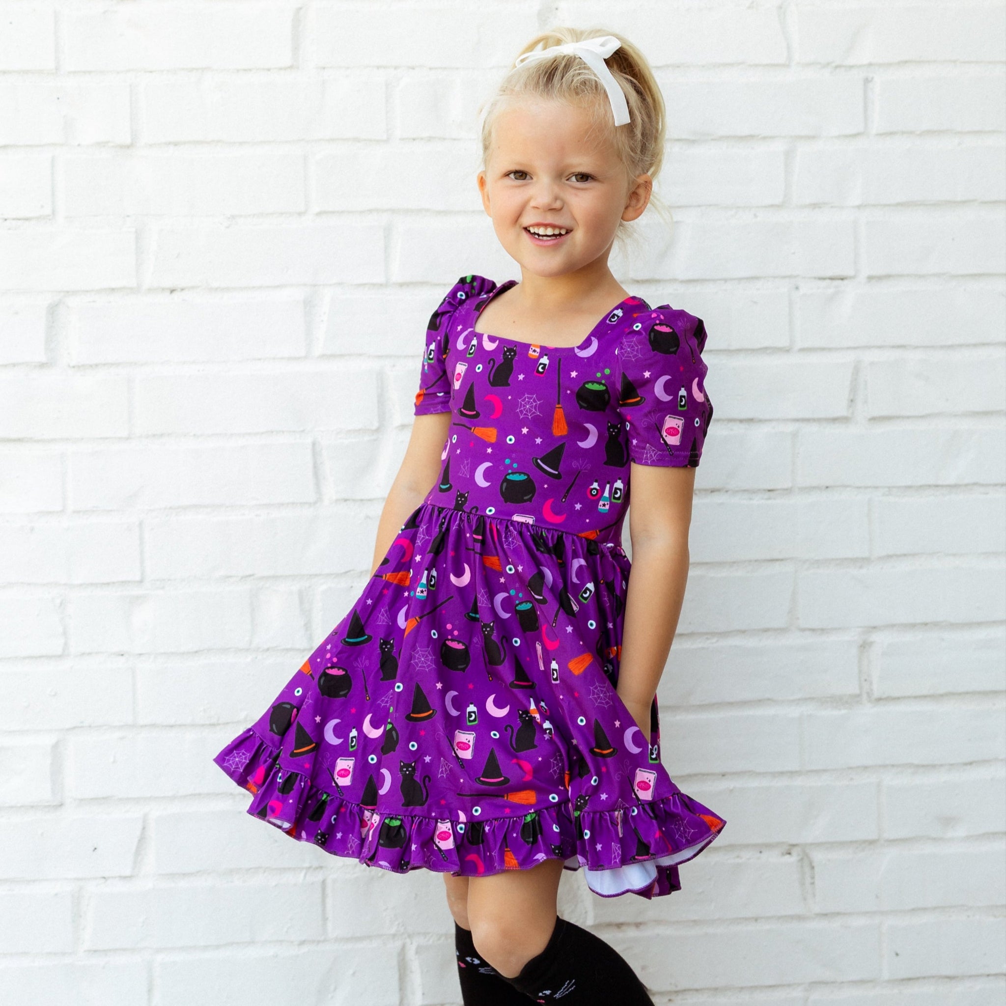 Young girl in a purple Halloween dress standing against a white brick wall with decorative bats.