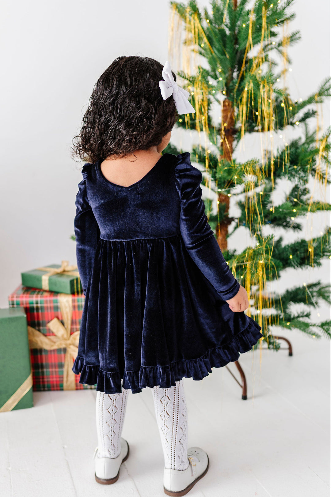 A toddler girl viewed from behind wearing a navy blue velvet long-sleeve dress, white tights, and white shoes, standing next to a decorated Christmas tree with wrapped gifts on a white floor.