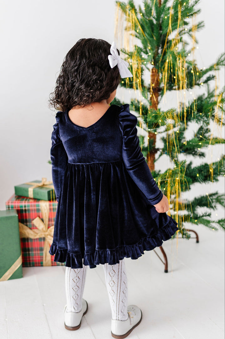 A toddler girl viewed from behind wearing a navy blue velvet long-sleeve dress, white tights, and white shoes, standing next to a decorated Christmas tree with wrapped gifts on a white floor.