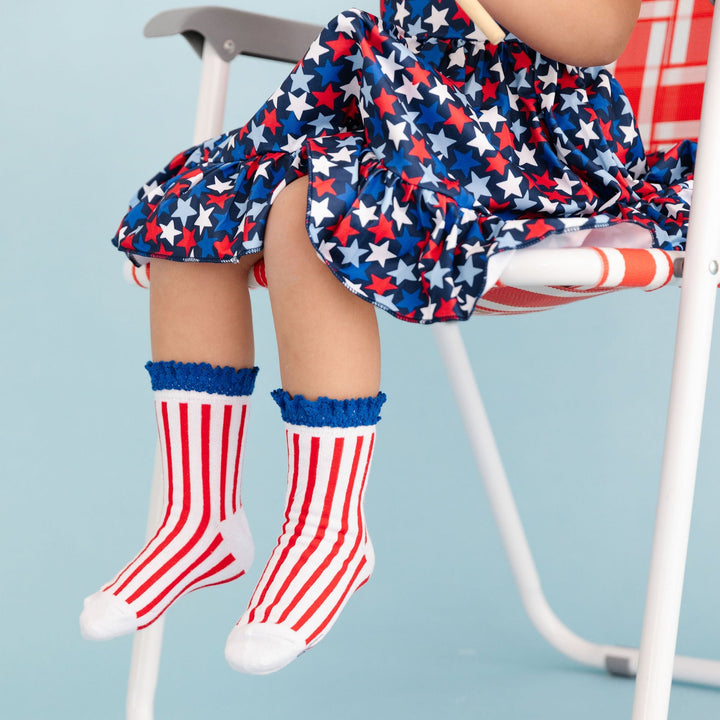 little girl sitting in yard chair wearing star print 4th of july dress with matching red, white and blue striped socks