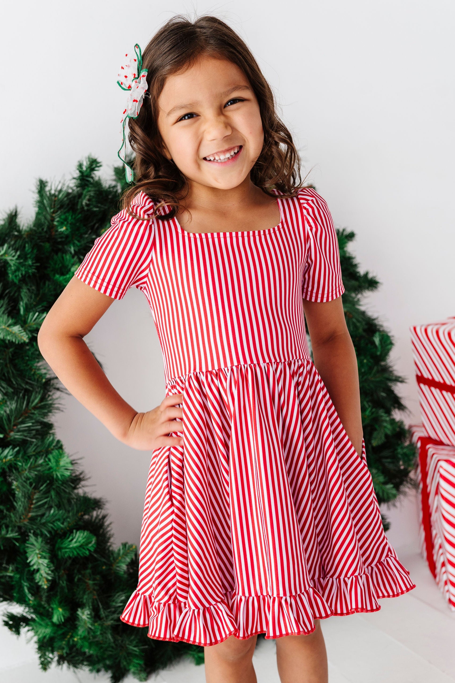 A young girl smiling and posing in a red and white vertically striped short-sleeve dress, standing in front of a green Christmas wreath and wrapped gifts.