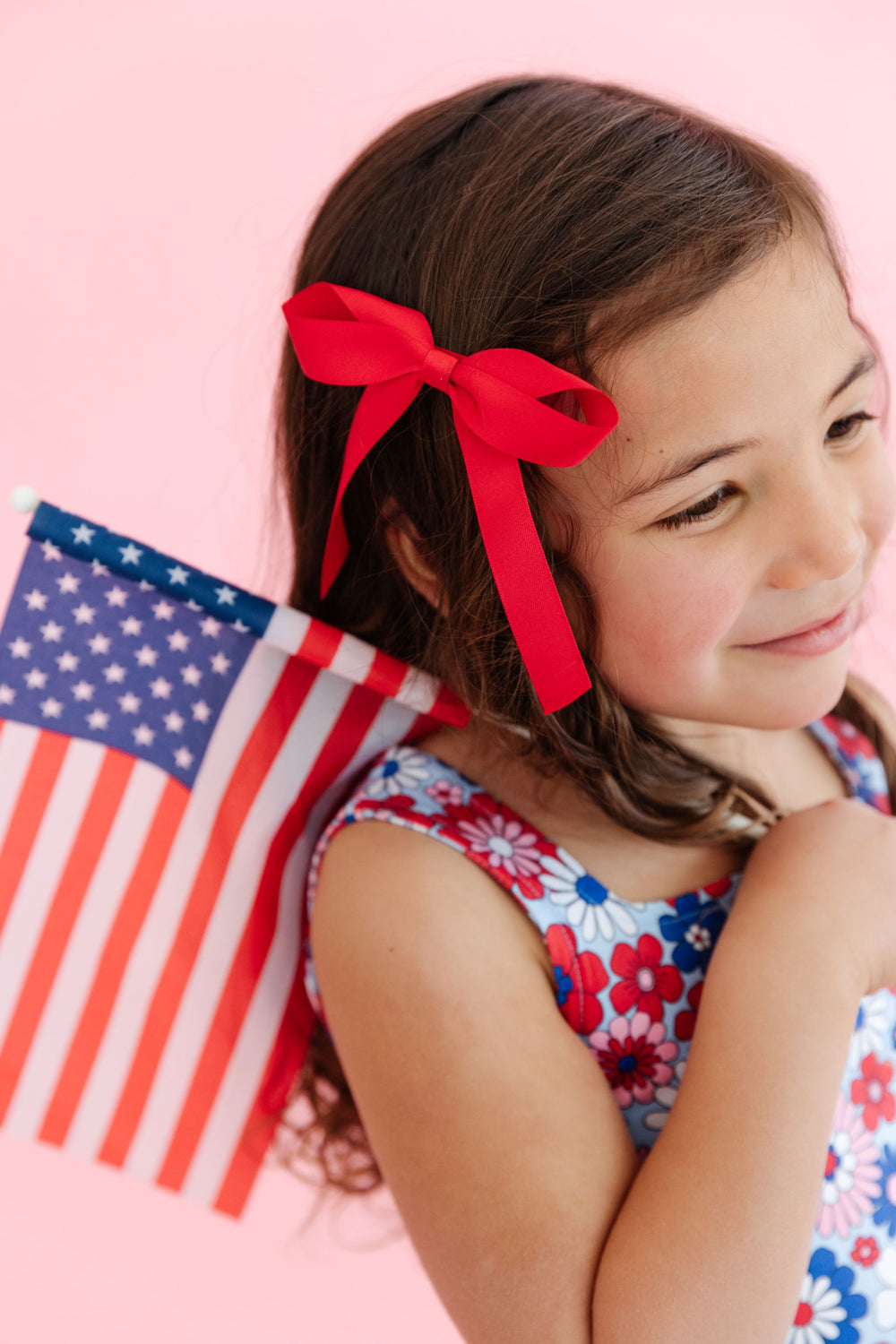 littler girl holding American flag with bright red satin bow on clip to match red, white and blue floral print dress