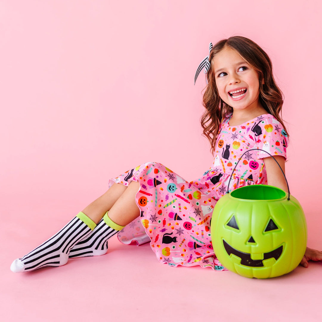 little girl sitting by green pumpkin bucket in pink v-neck twirl dress with colorful Halloween themed print paired with black and white striped ankle socks with sparkly green trim