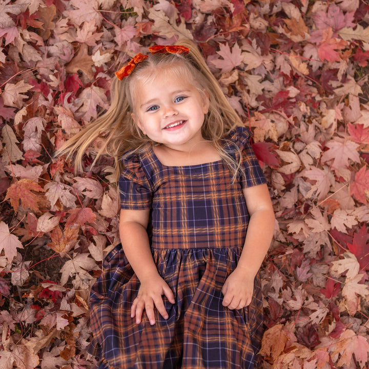 little girl laying in a fall leaf pile in brown and black plaid dress with matching pumpkin spice velvet pigtail bows