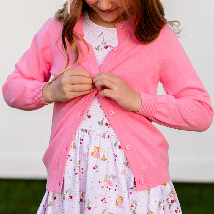 girl buttoning taffy pink cotton knit cardigan over lavender gingham twirl dress with vintage kitten print