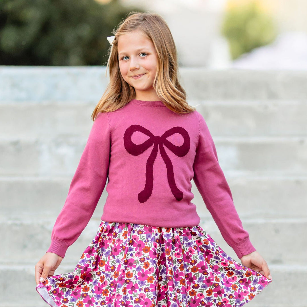 Young girl in a pink sweater with a bow and floral skirt standing on steps.