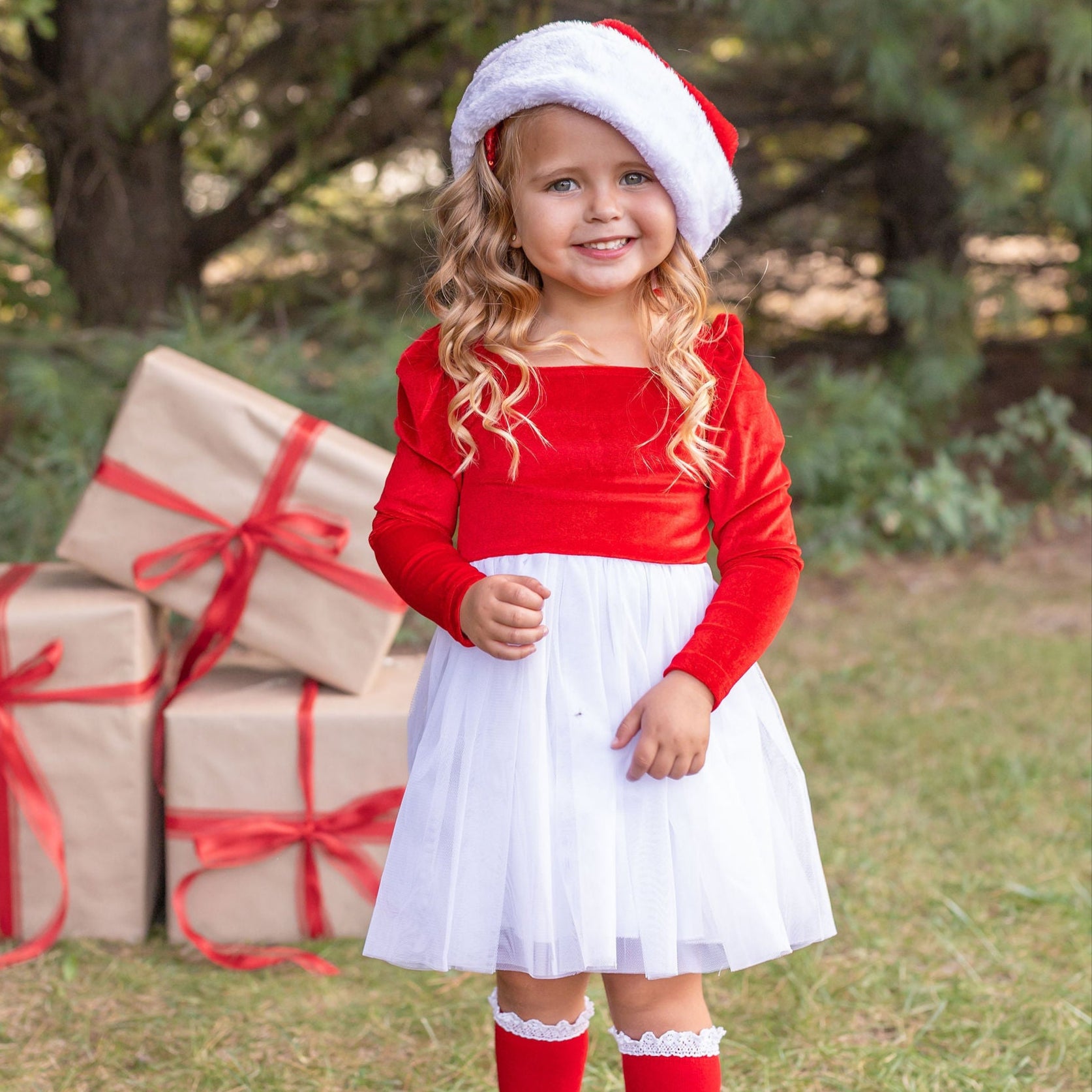 Child in Christmas outfit with red sweater, white skirt, and Santa hat, standing next to wrapped gifts outdoors.