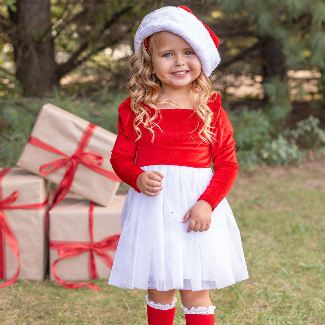 Child in Christmas outfit with red sweater, white skirt, and Santa hat, standing next to wrapped gifts outdoors.