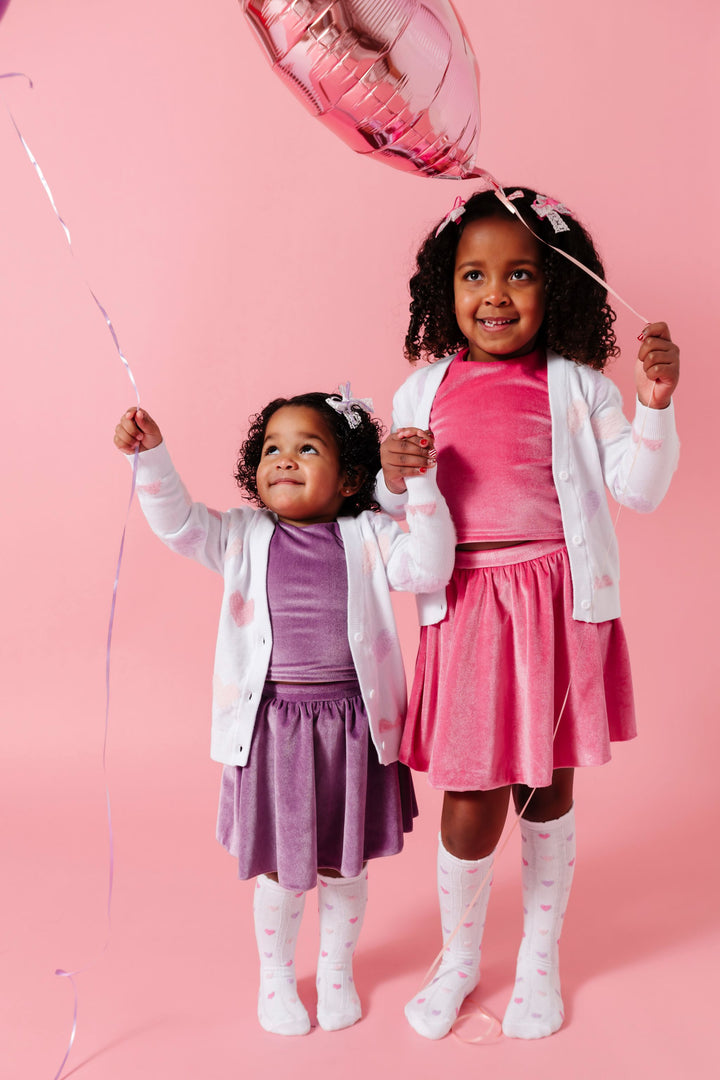 sisters holding heart balloons in pink and lavender velvet skort and top sets paired with white V-neck cardigan with pastel fuzzy hearts and matching white scallop trimmed knee socks with hearts