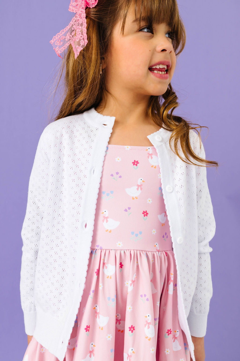 little girl wearing pink dress with playful spring goose and flower print and white pointelle knit cardigan over the top
