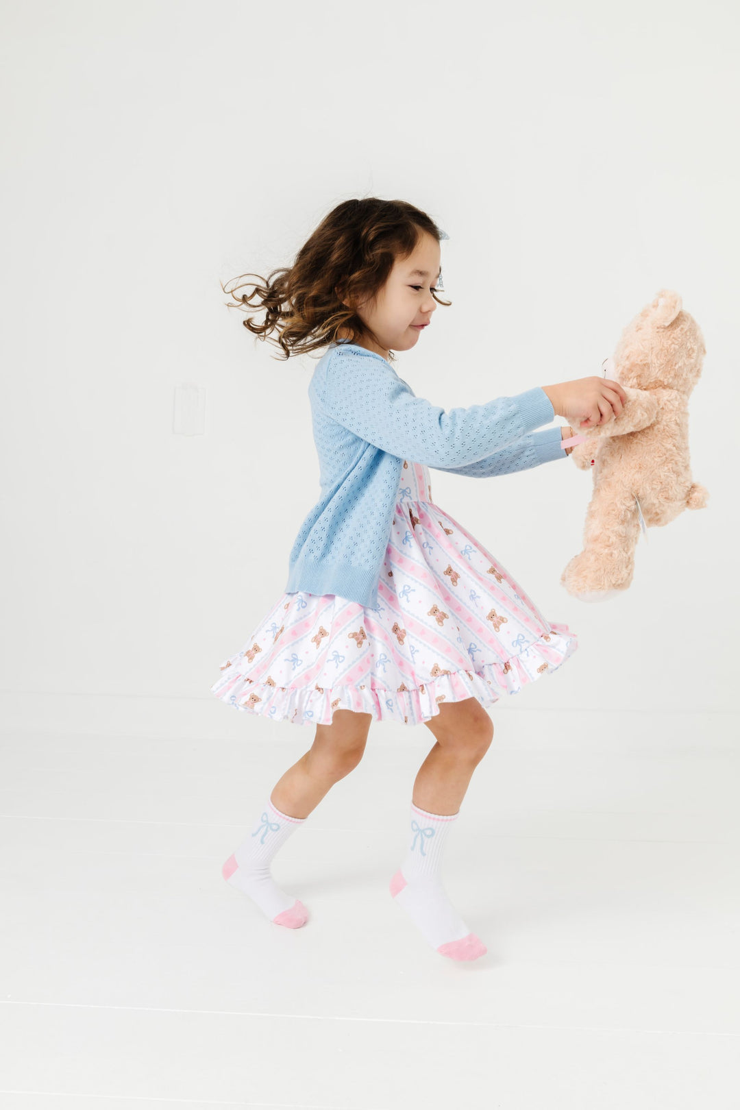 little girl twiring with teddy bear in teddy print twirl dress and matching blue pointelle sweater and blue bow print crew socks