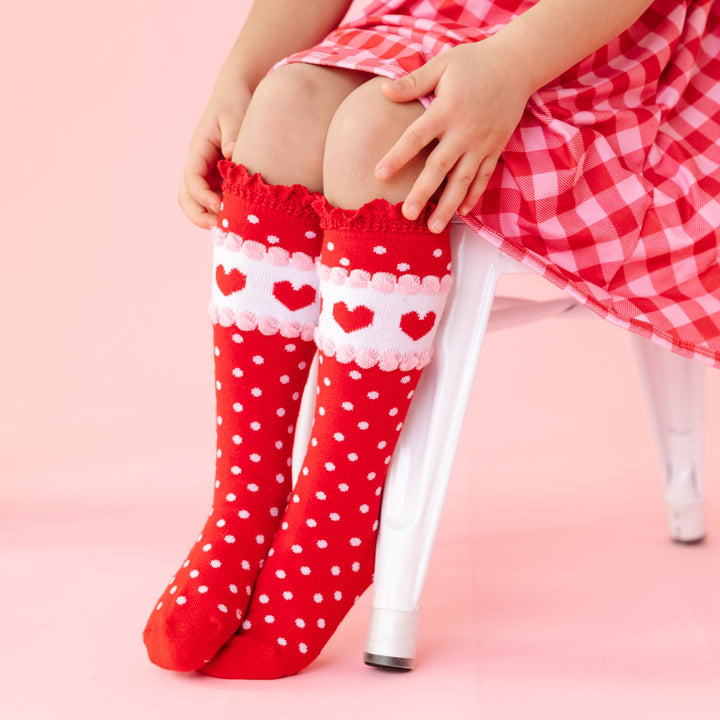 little girl in Valentine's inspired red lace top knee high socks with polka dots and heart detail paired with matching red and pink gingham dress