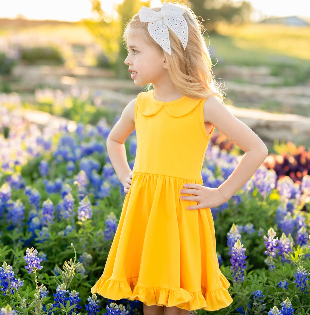 little girl standing in field of flowers in bright yellow tank top style summer dress with white bow in her hair