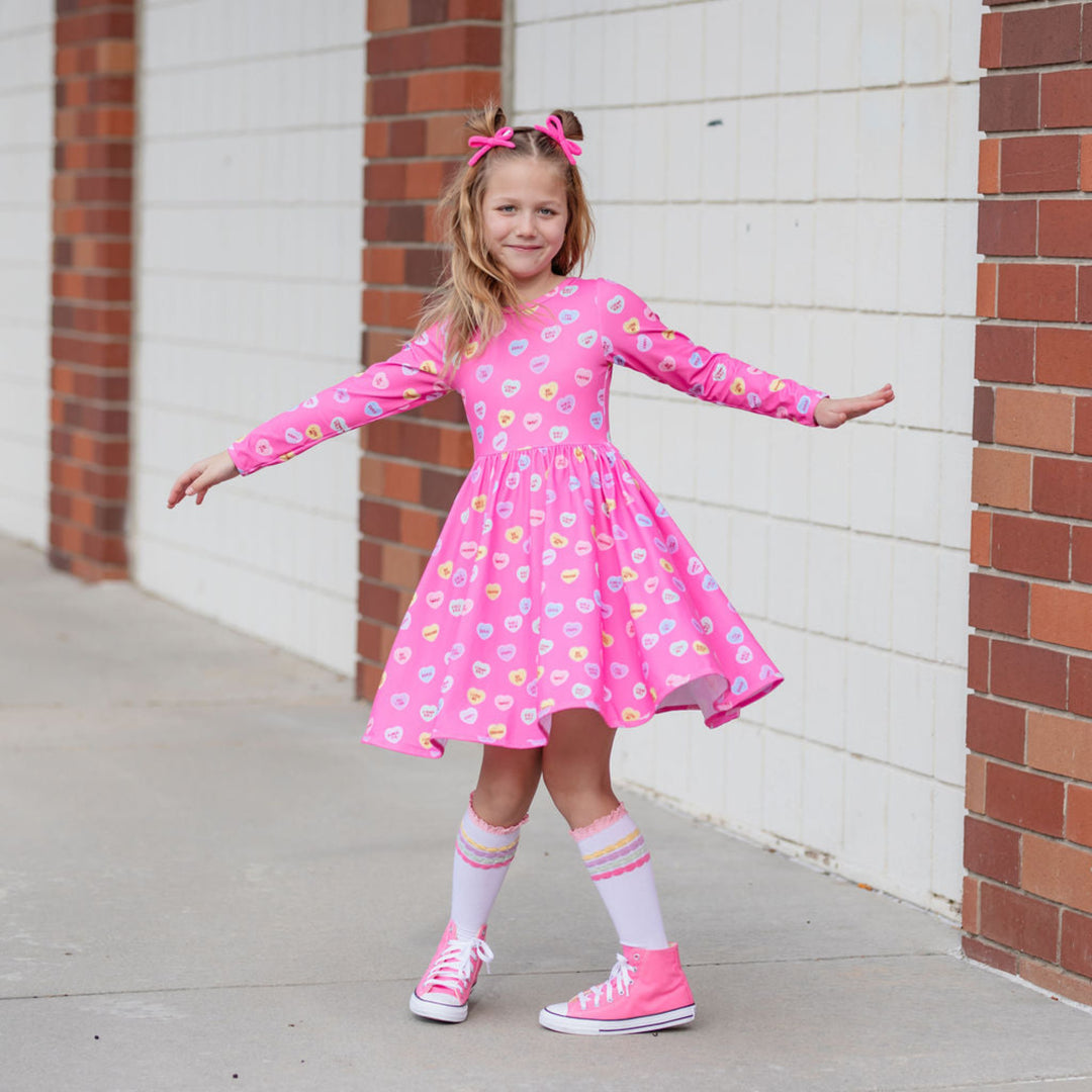 little girl in long sleeve pink Valentines twirl dress with conversation heart print, matching pastel striped knee highs and pink yarn pigtail bows