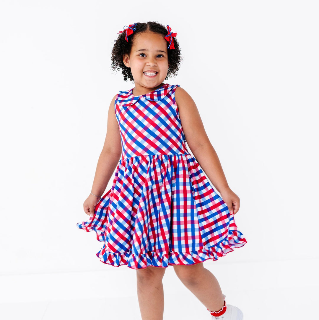 little girl wearing red, white sand blue gingham print twirl dress with peter pan collar and matching socks and pigtail bows