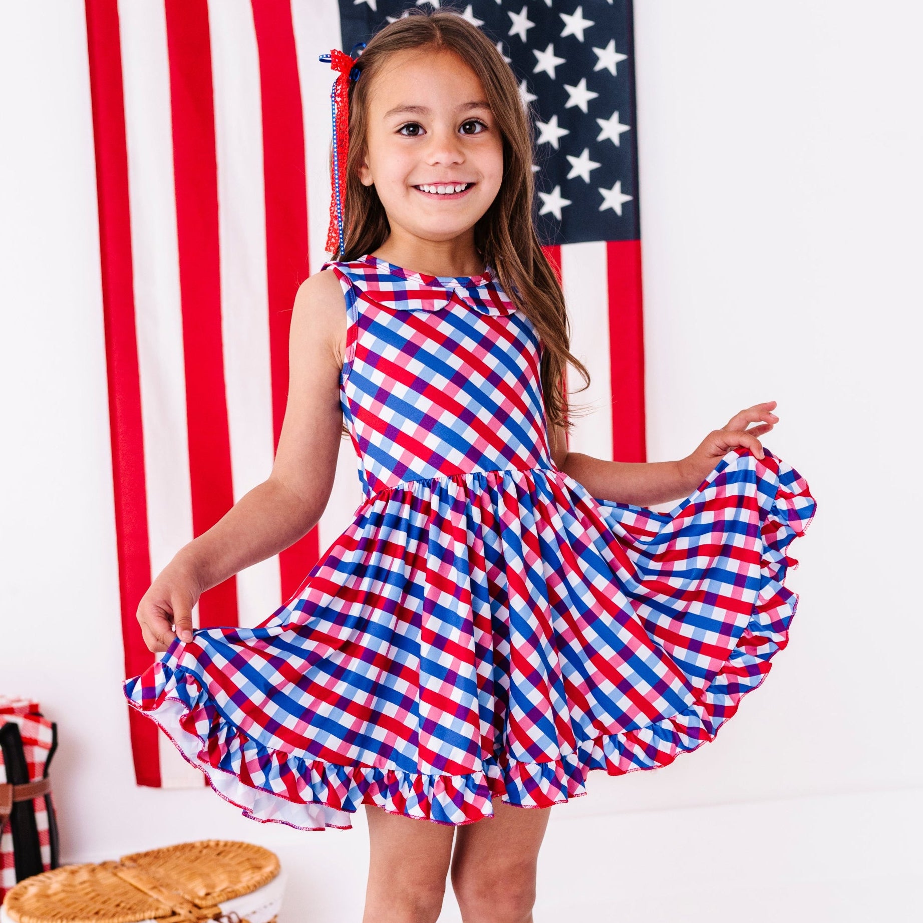 little girl with picnic accessories in front of American flag with red, white and blue gingham sleeveless collared twirl dress for the 4th of July 