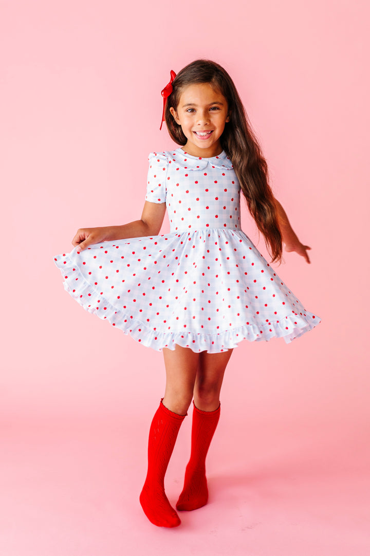girl twirling in blue and white apple print gingham twirl dress paired with bright red pointelle knee socks and red satin hair bow