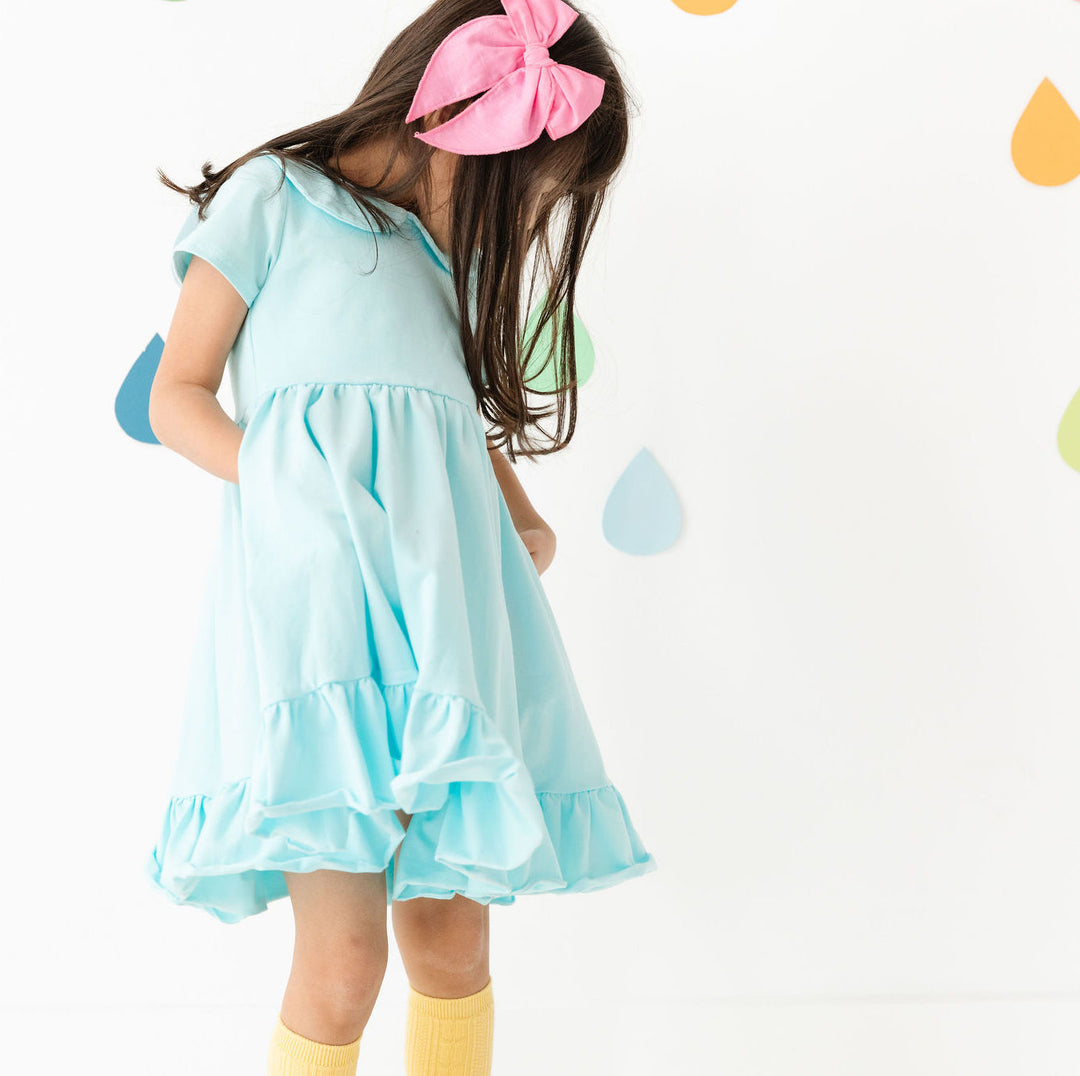 little girl wearing aqua blue party dress with pockets and bright pink bow in her hair