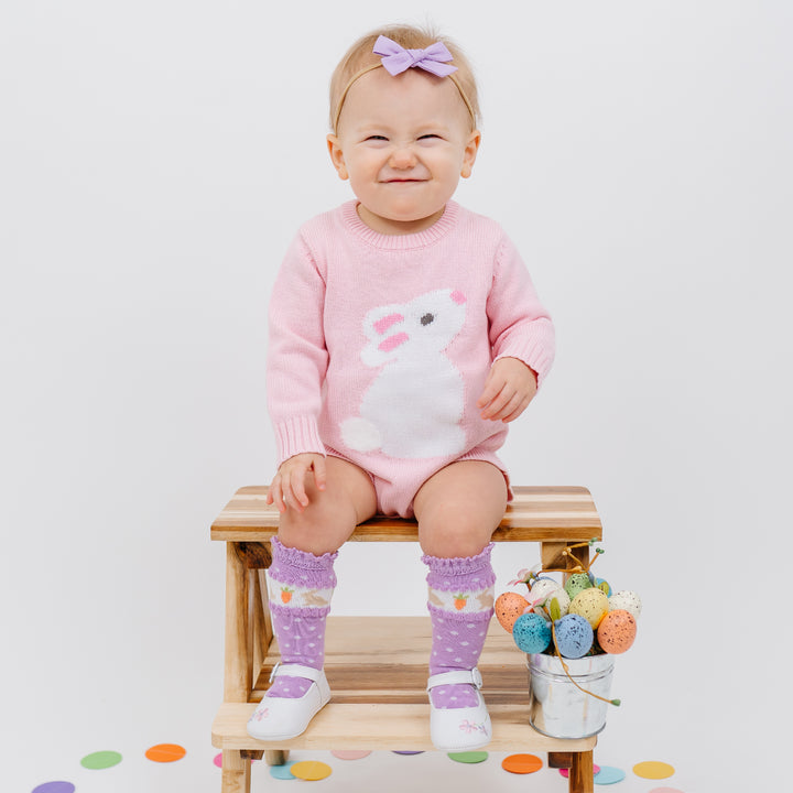 baby in an easter pink sweater romper with a white bunny, also wearing a purple bow and purple knee high socks