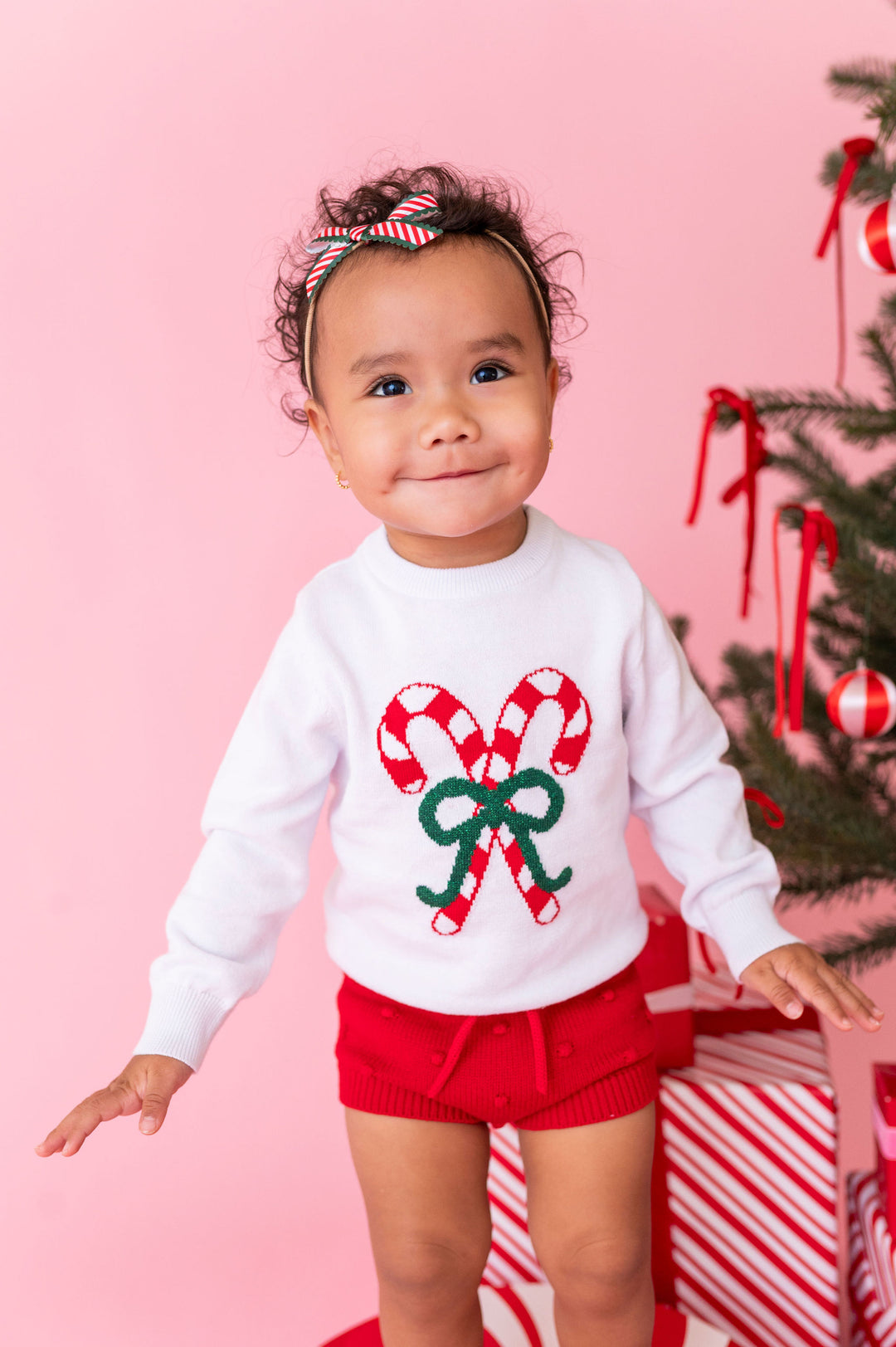 A toddler wearing a white long-sleeve sweater with a red and green candy cane and bow graphic, paired with red knit shorts, standing against a pink background next to a decorated Christmas tree.
