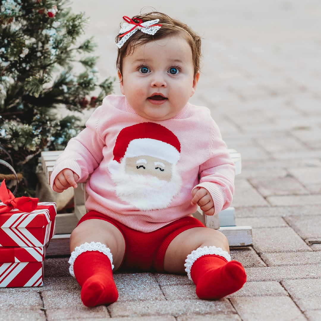 baby girl sitting by Christmas tree in pink Santa sweater with red bloomers and red and white socks and bow