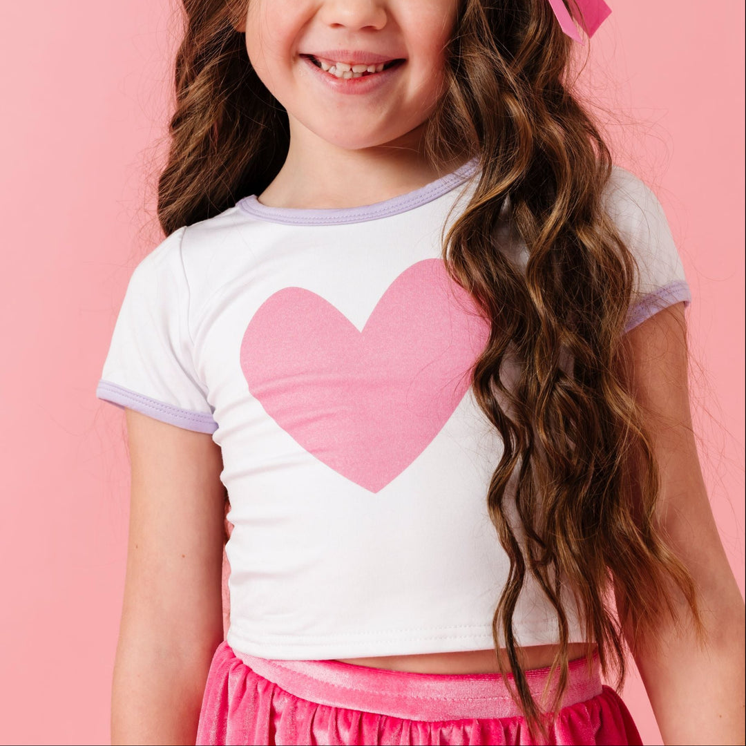 young girl in white bamboo stretchy fitted top with pink heart design and lavender trim paired with pink satin hair bow and matching pink velvet skort