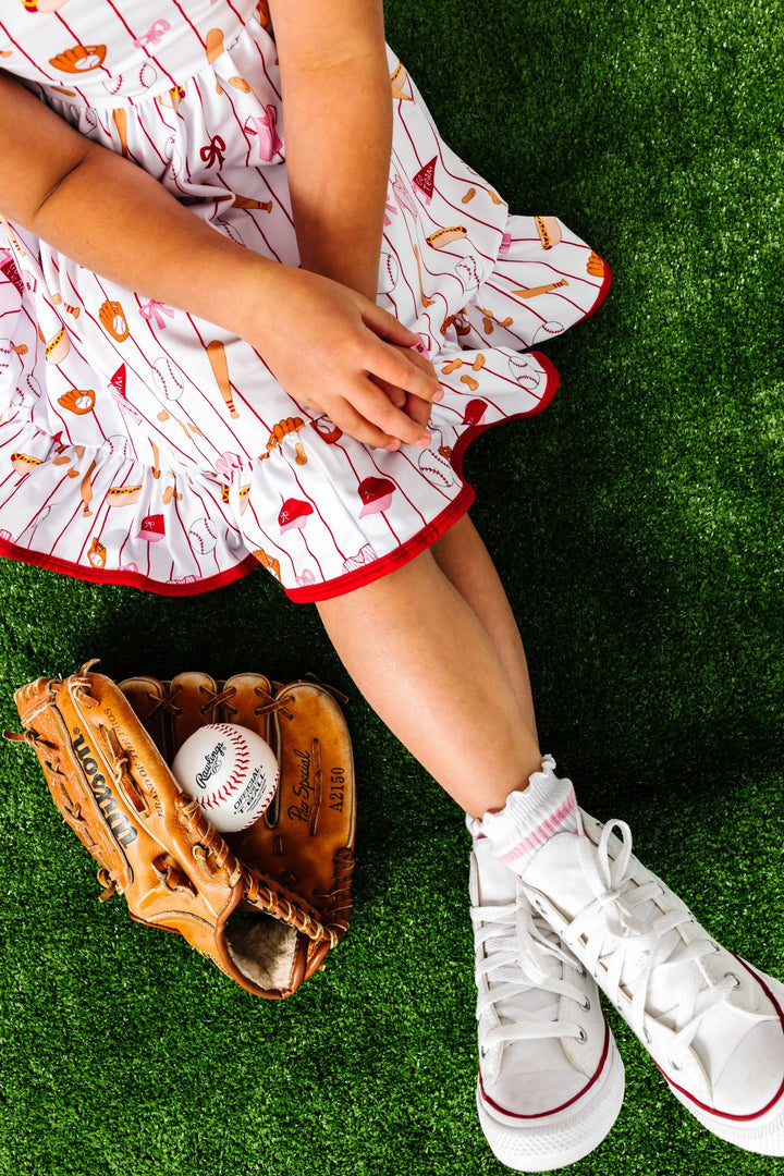 little girl sitting in grass in baseball print twirl dress paired with lettuce trim pink striped midi socks and white high tops beside glove and baseball