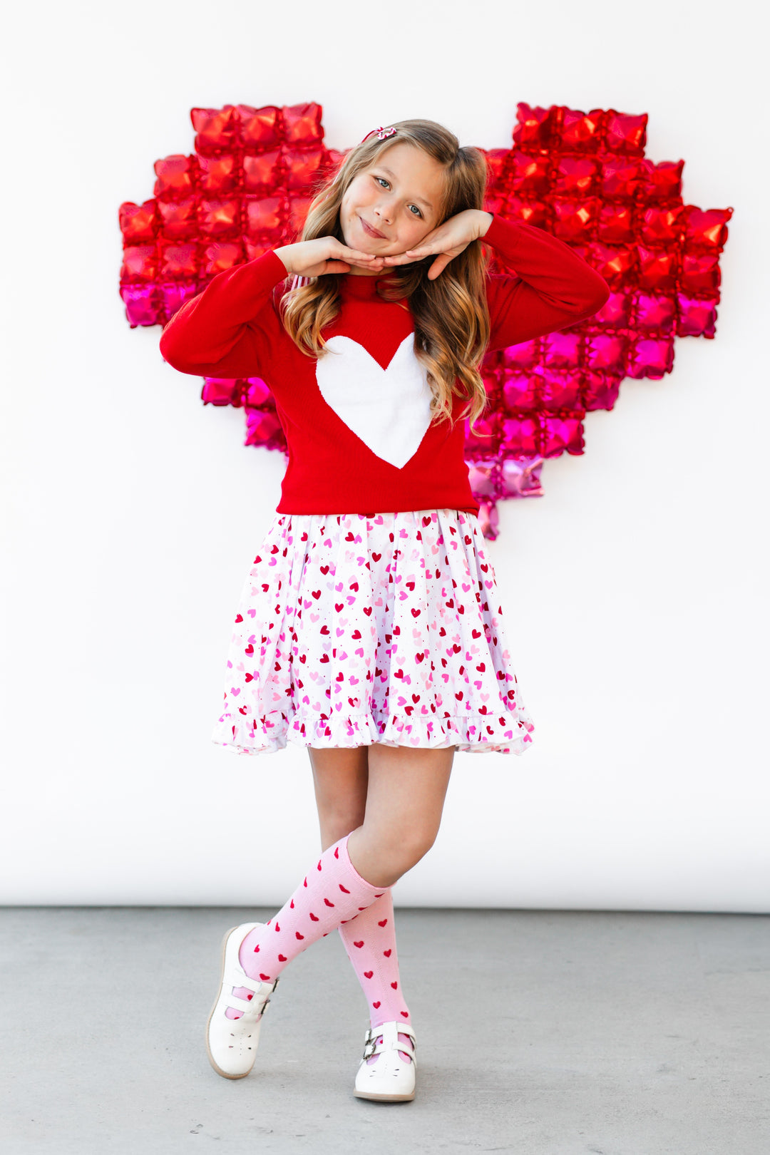 girl posing in front of heart balloon wearing bright red cotton knit valentines sweater with white heart design paired with white twirl dress with confetti heart print and pink knee socks with red hearts
