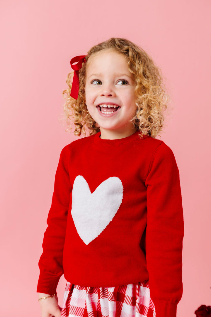 girl in bright red sweater with single white heart design for Valentine's Day paired with red and pink plaid dress